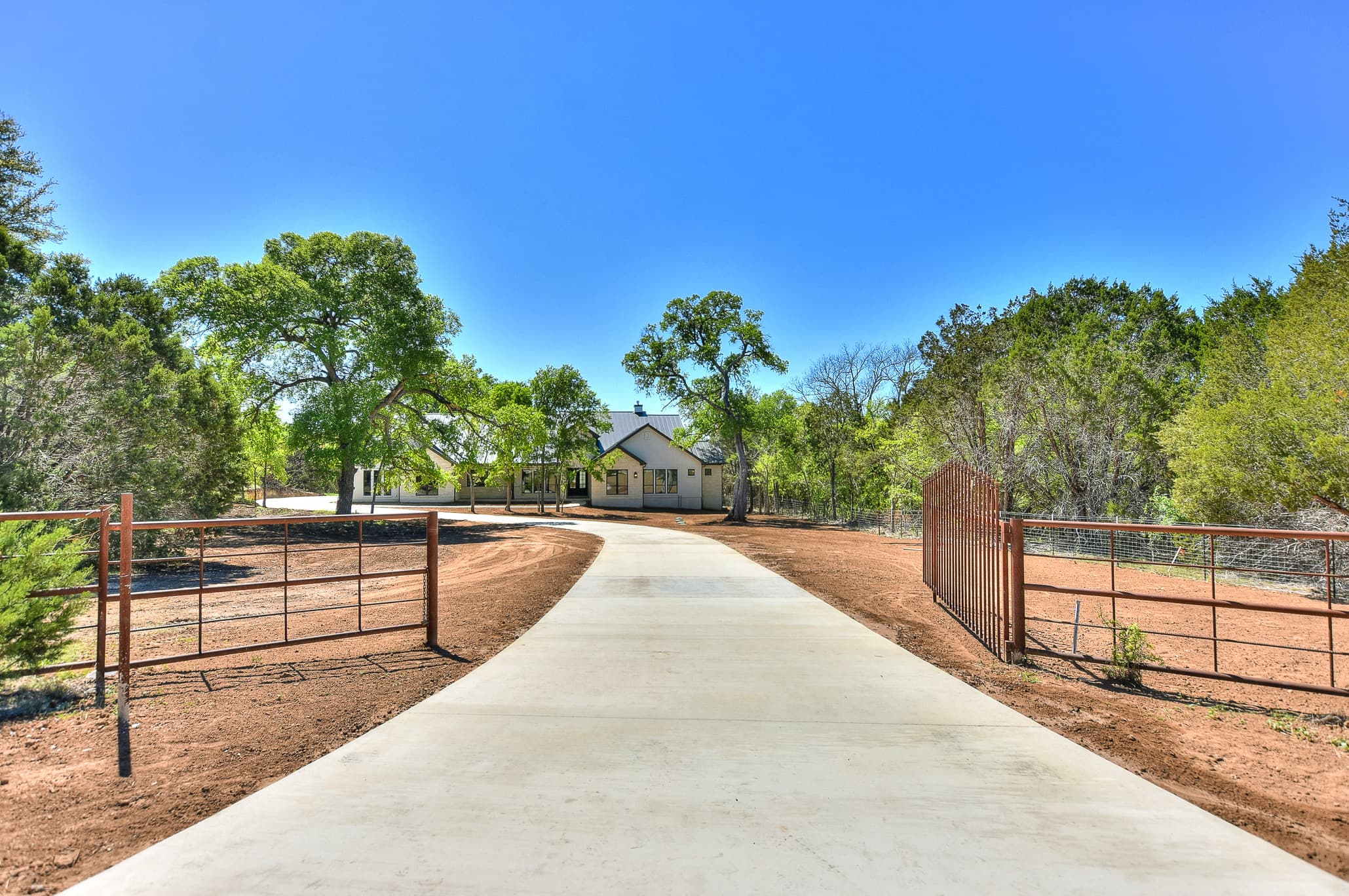 Long concrete driveway leading to the house past ranch-style rust-colored gates and oak trees