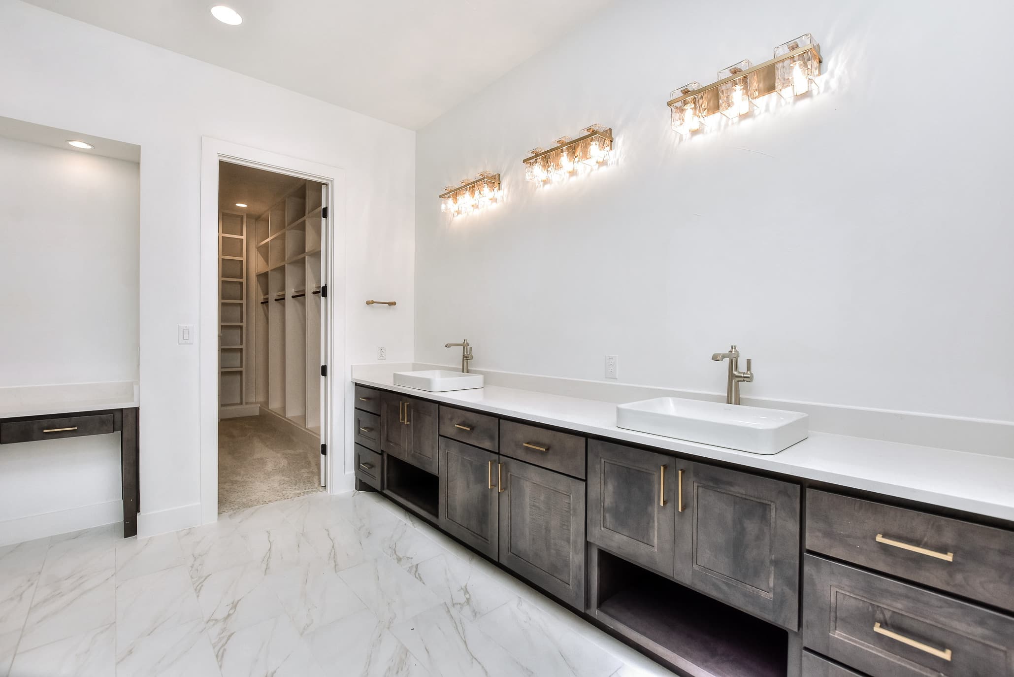 Primary bath with a dual vanity in gray wood, vessel sinks, brass vanity lights and marble-look floor
