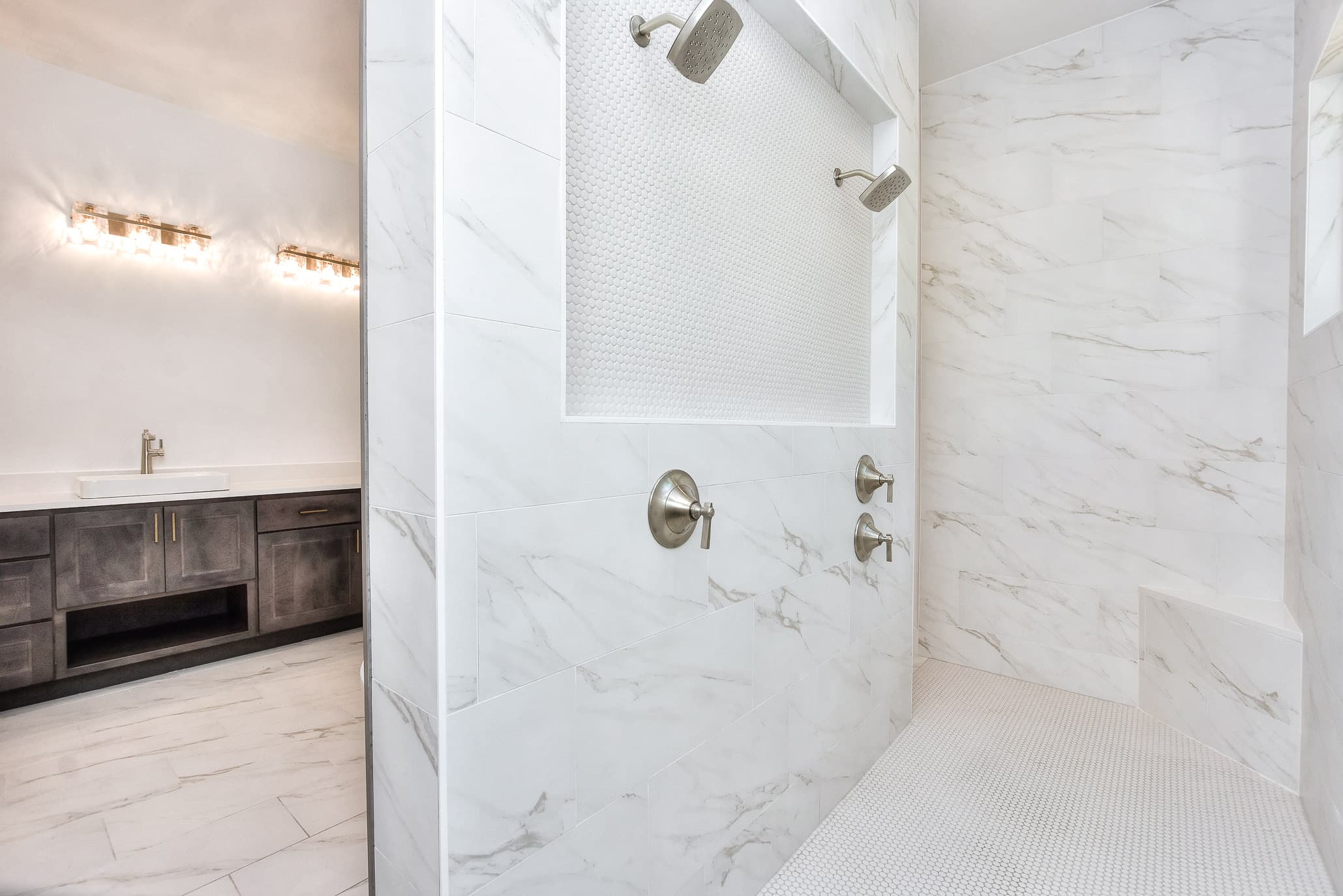 Marble-look walk-in shower with a recessed niche beside a gray wood vanity under brass lights