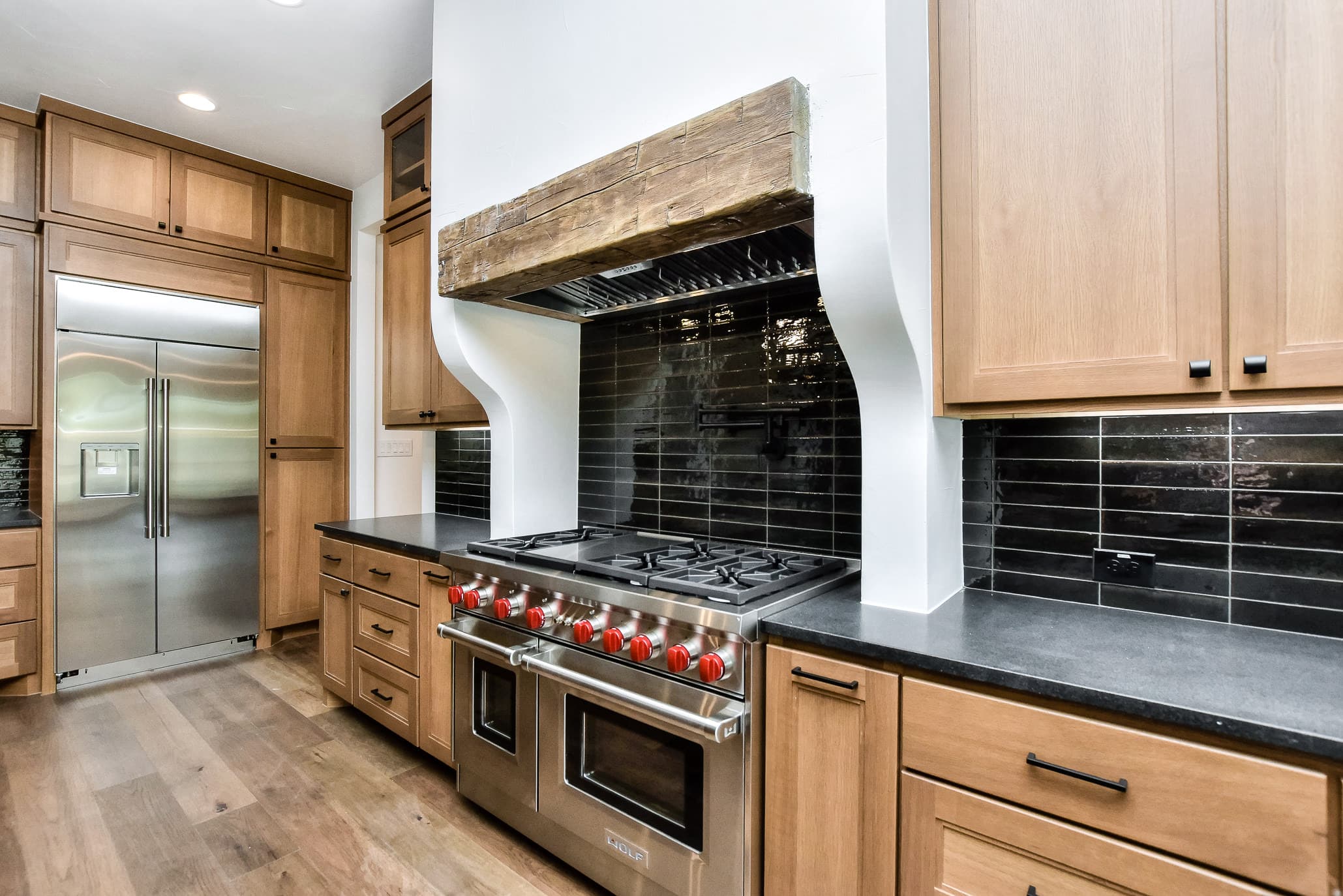 Professional range with red knobs under a plastered hood with a reclaimed wood mantel and black tile backsplash