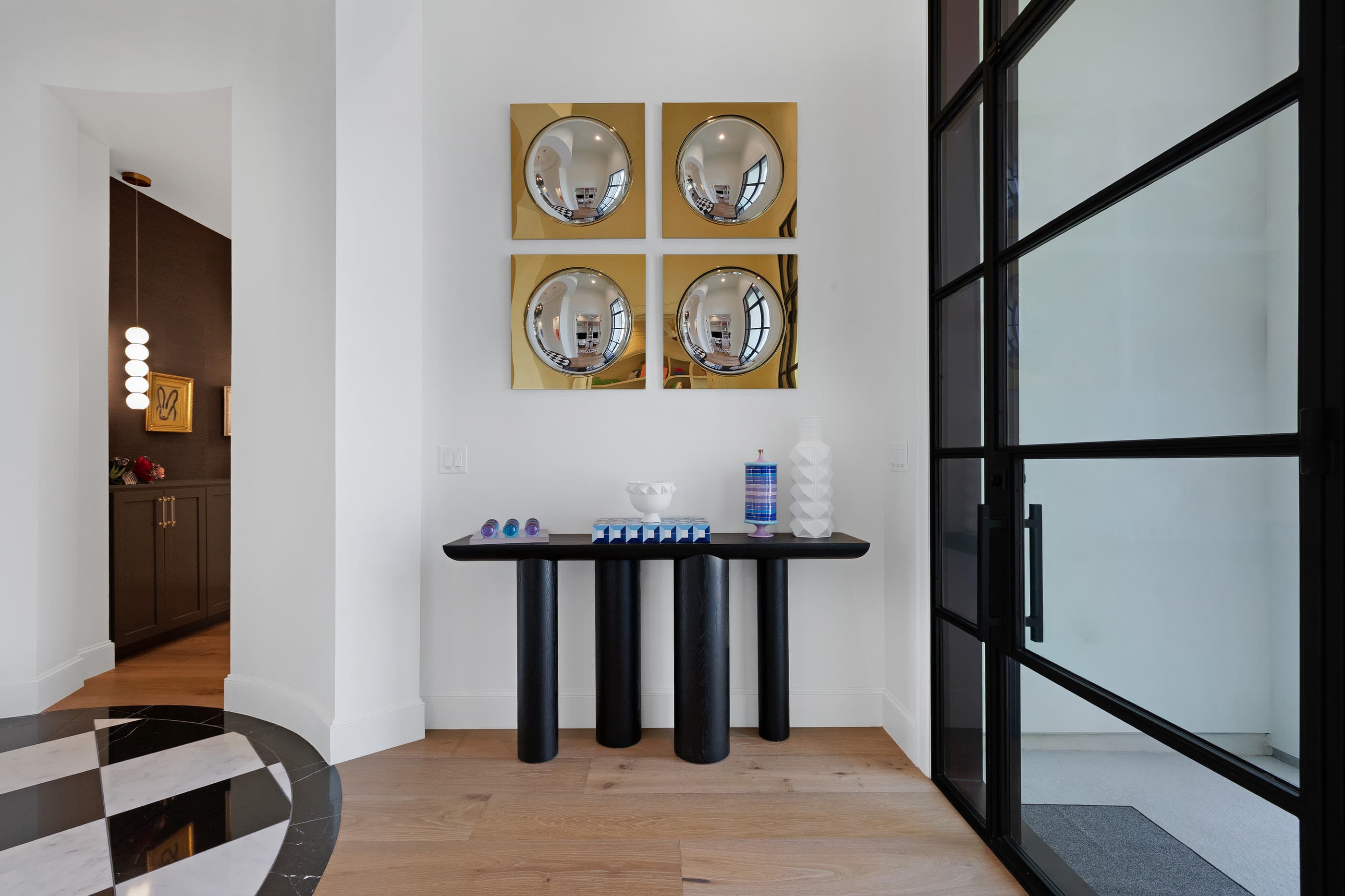 Front entry console table beneath gold convex mirrors beside a steel-framed glass door and checkerboard floor inlay