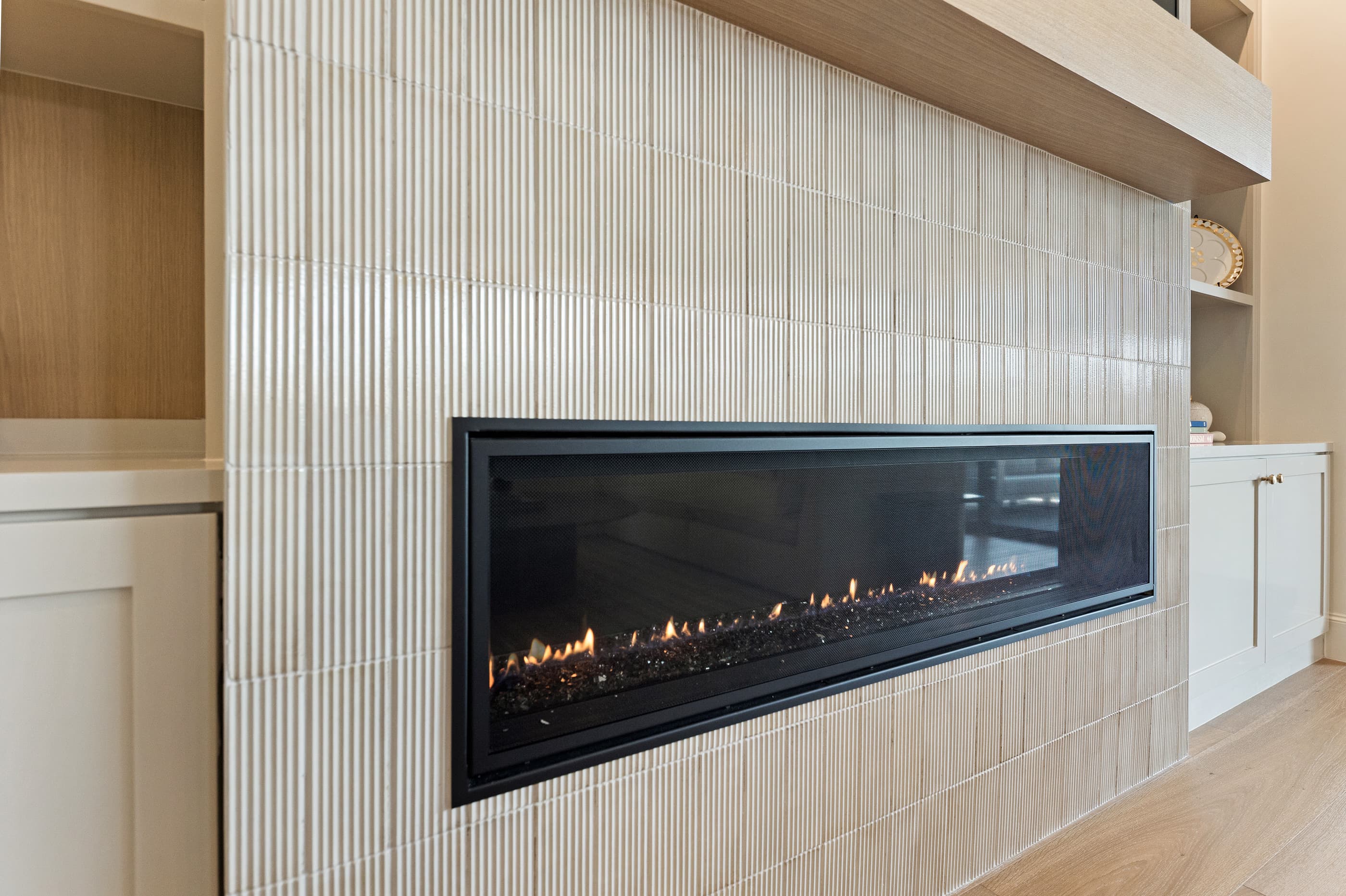 Linear gas fireplace surrounded by fluted vertical tile with a floating oak mantel