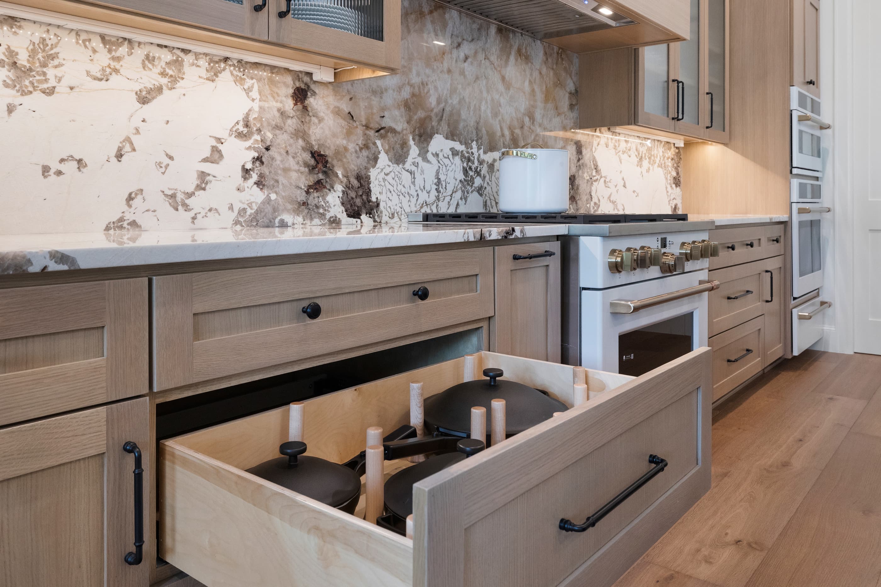 Pot-and-pan drawer open under a white range with stone backsplash in a custom oak kitchen