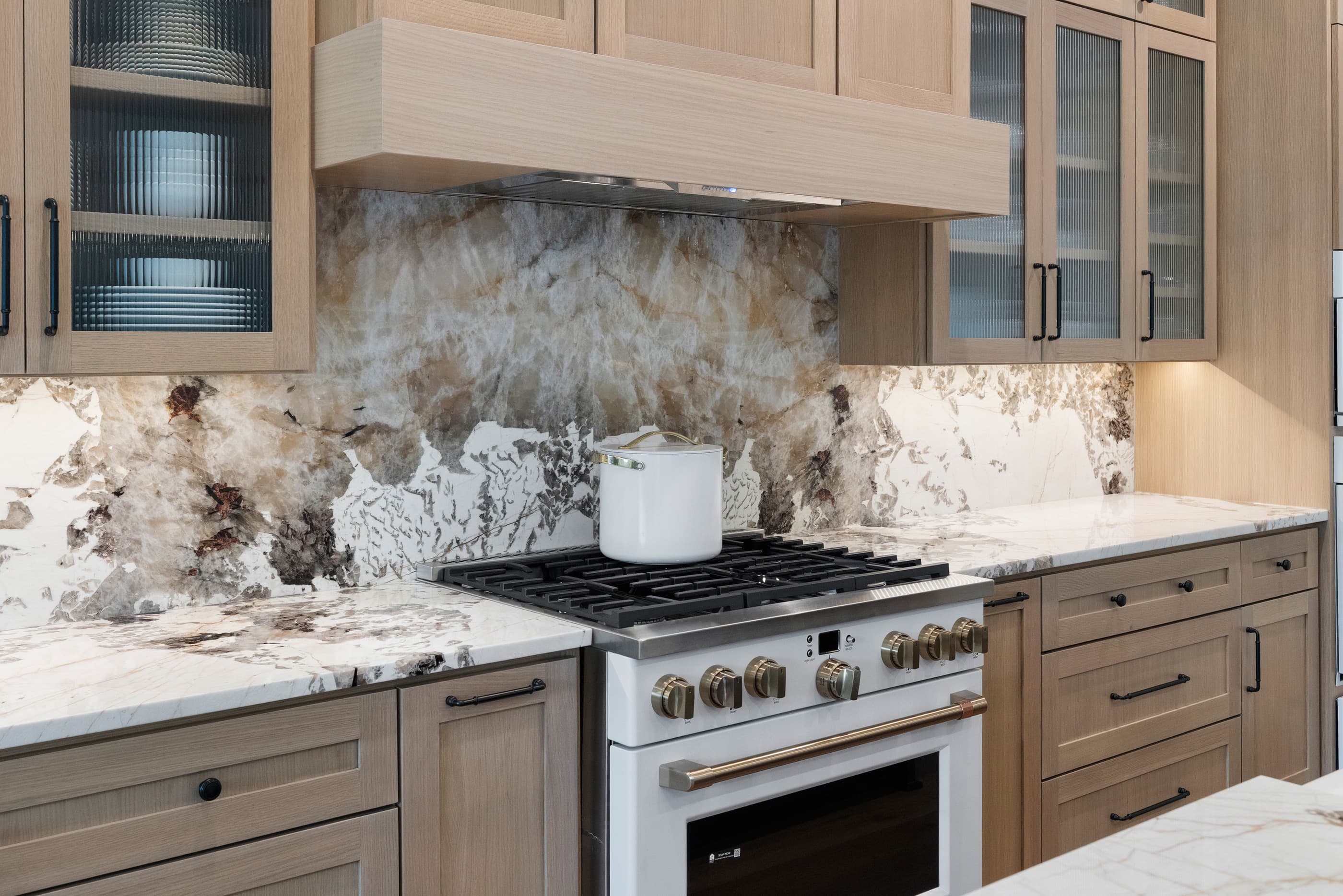 Gas range under an oak hood with natural stone slab backsplash and reeded-glass cabinets