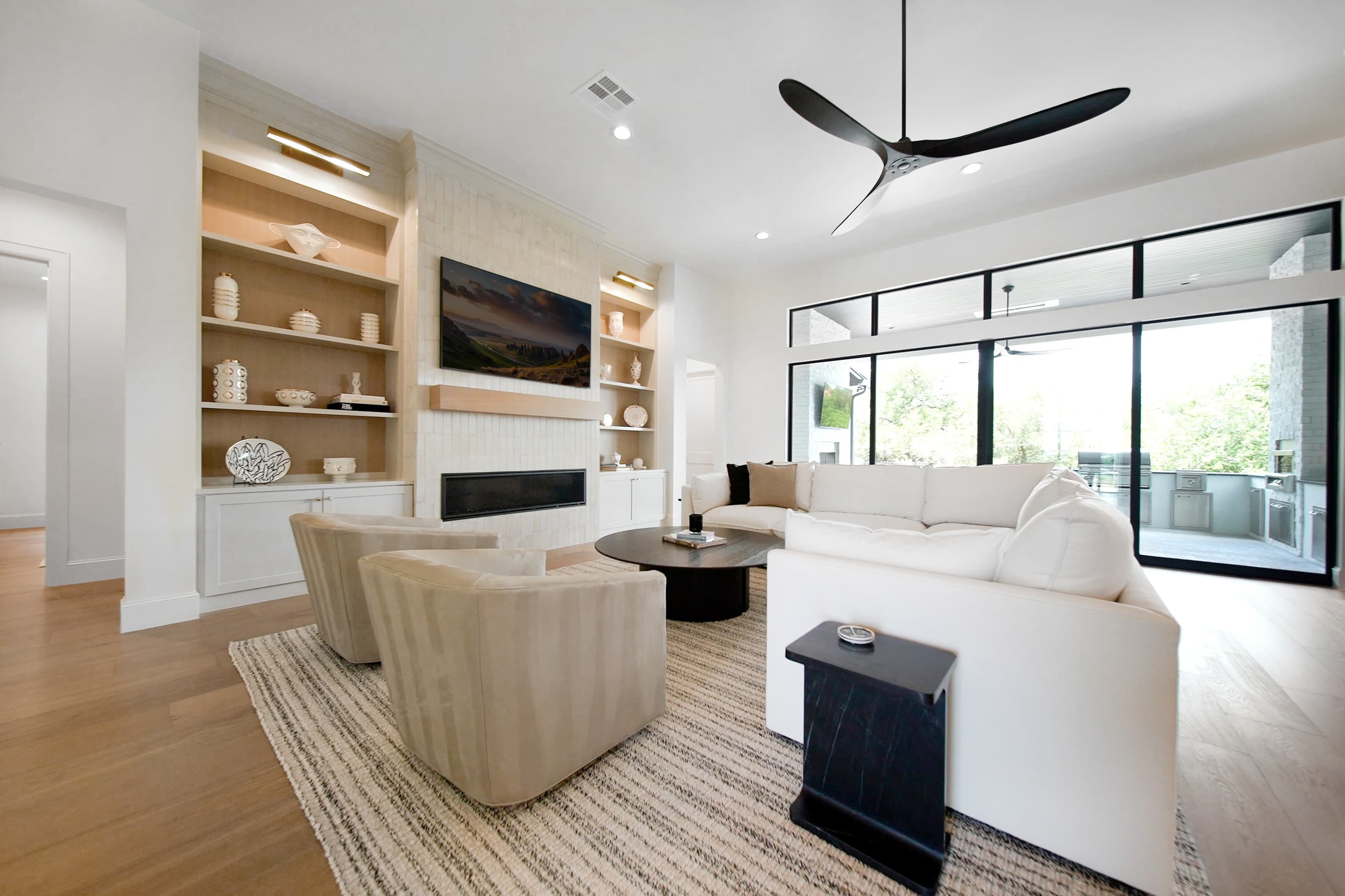 Open great room with white sectional, built-in shelving, ceiling fan and sliding glass walls to the patio