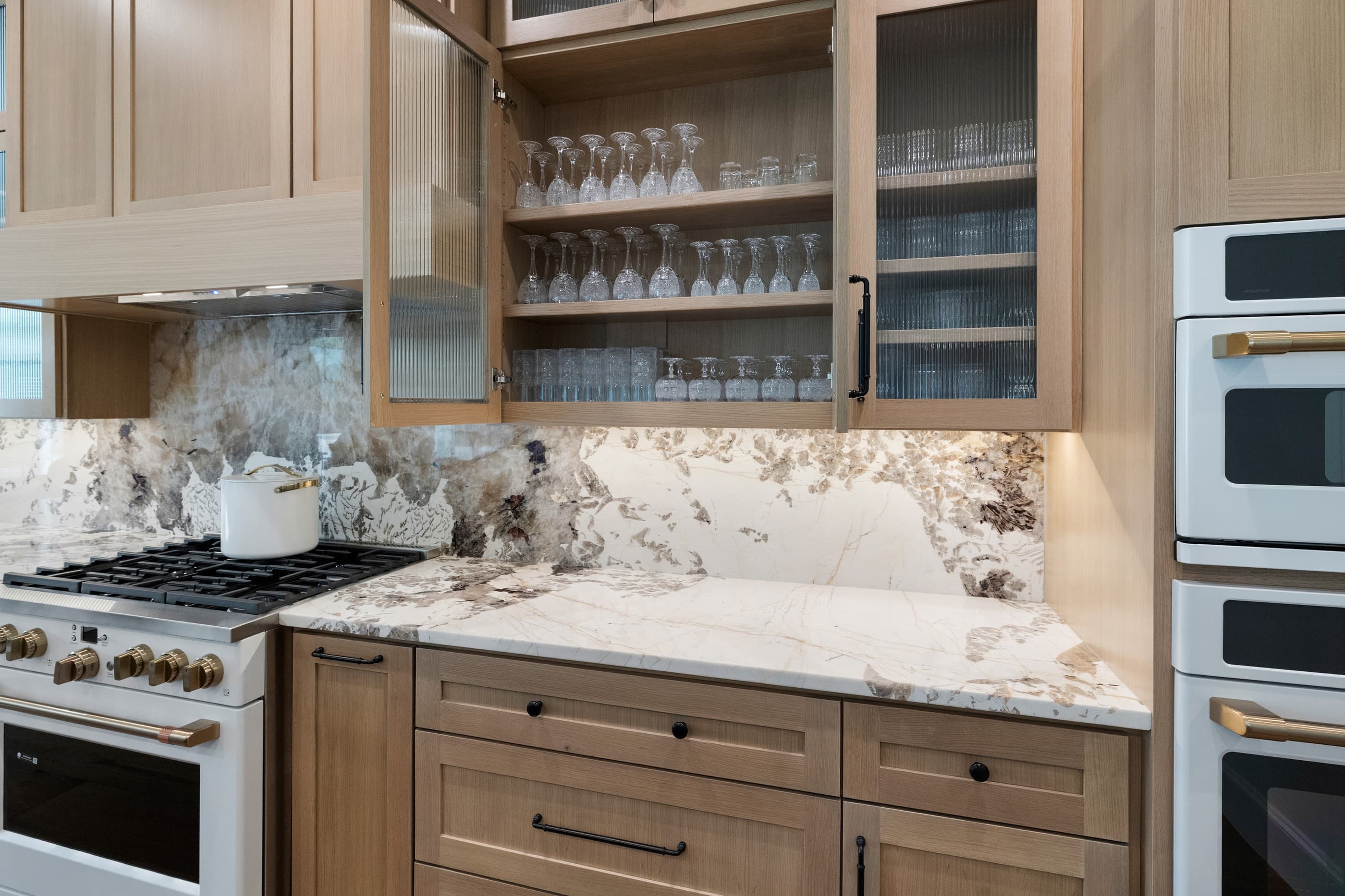 Kitchen glass-front cabinets with stemware beside built-in ovens and a white gas range