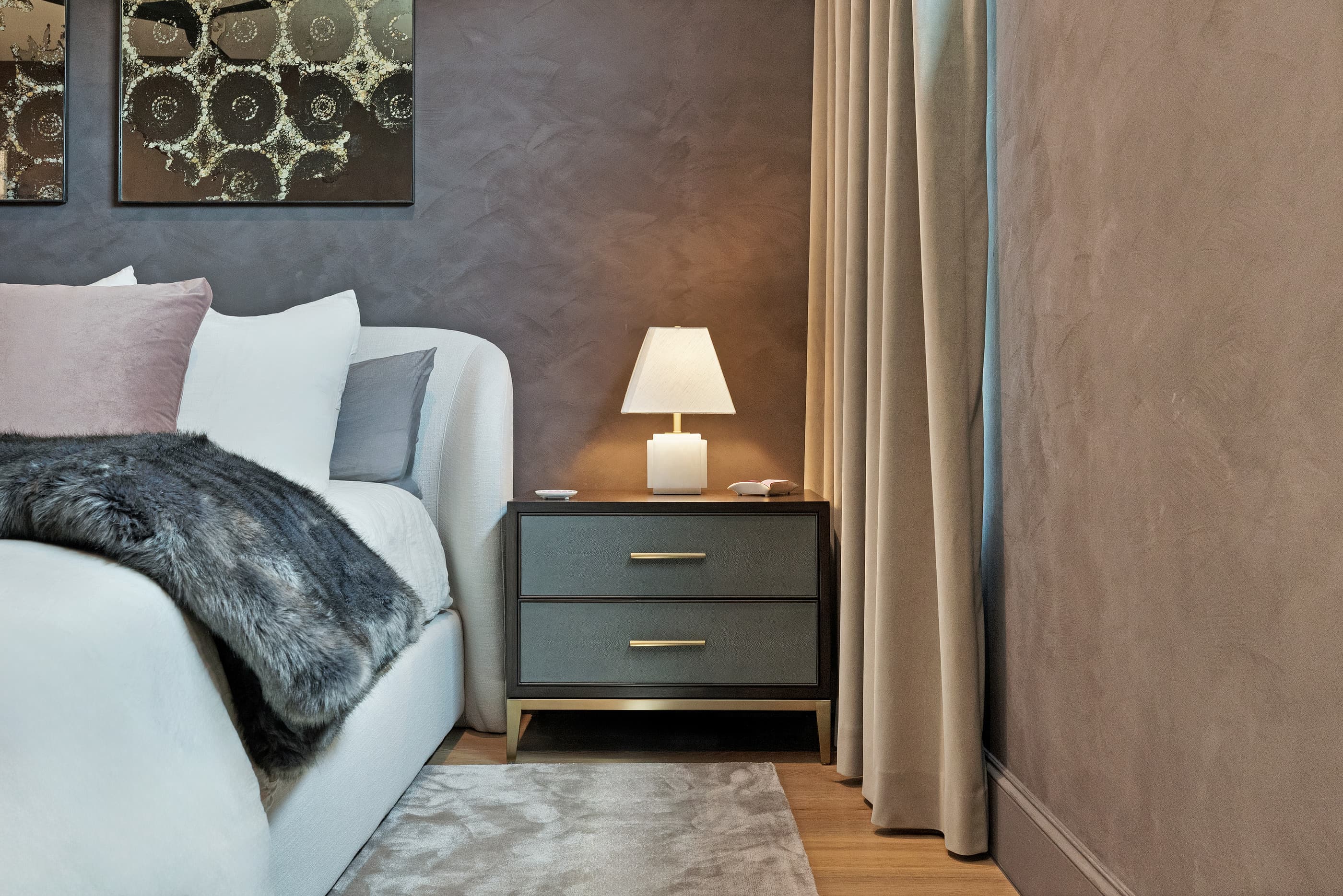 Bedside table and lamp beside an upholstered bed against a plaster-finish wall with floor-length drapes