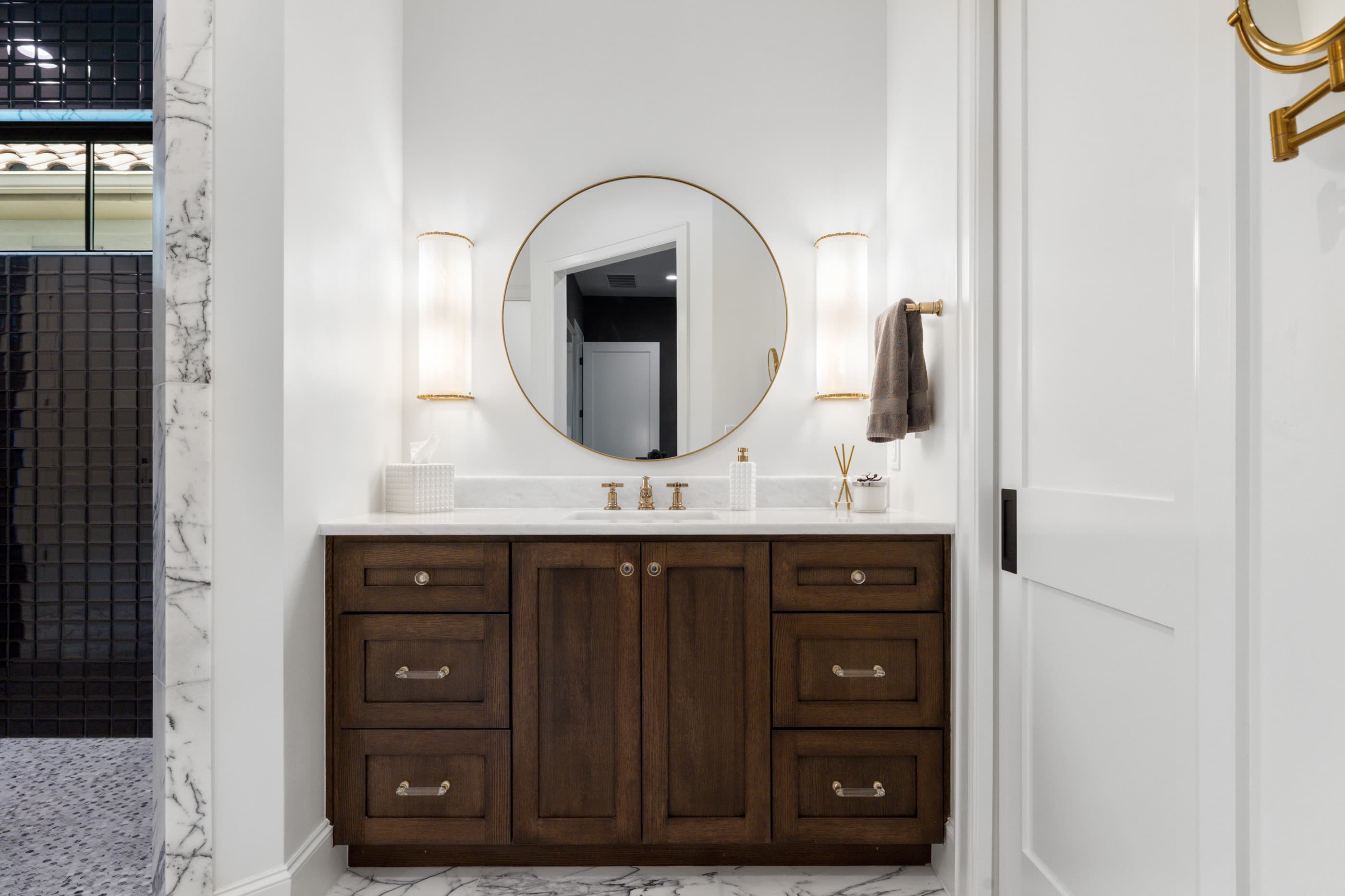 Primary bathroom vanity in dark stained wood with a round mirror, brass fixtures and marble accents