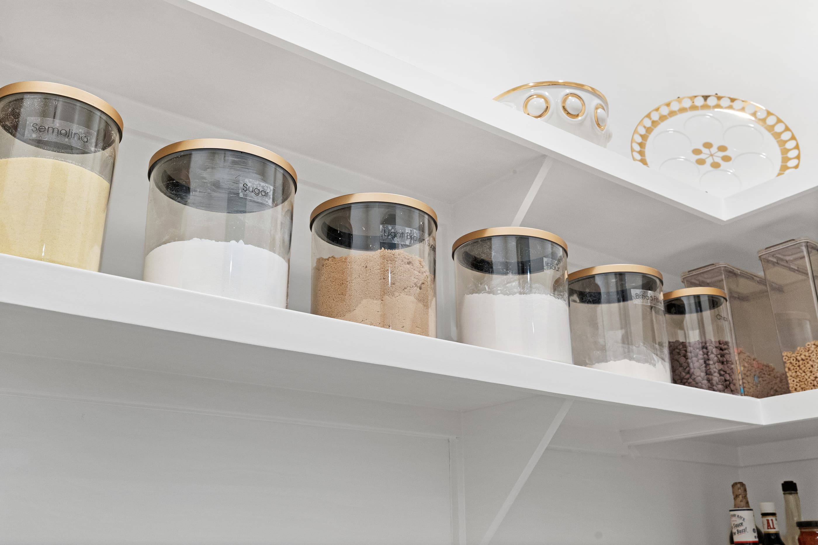 Pantry shelf of glass canisters with gold lids holding flour, sugar and grains