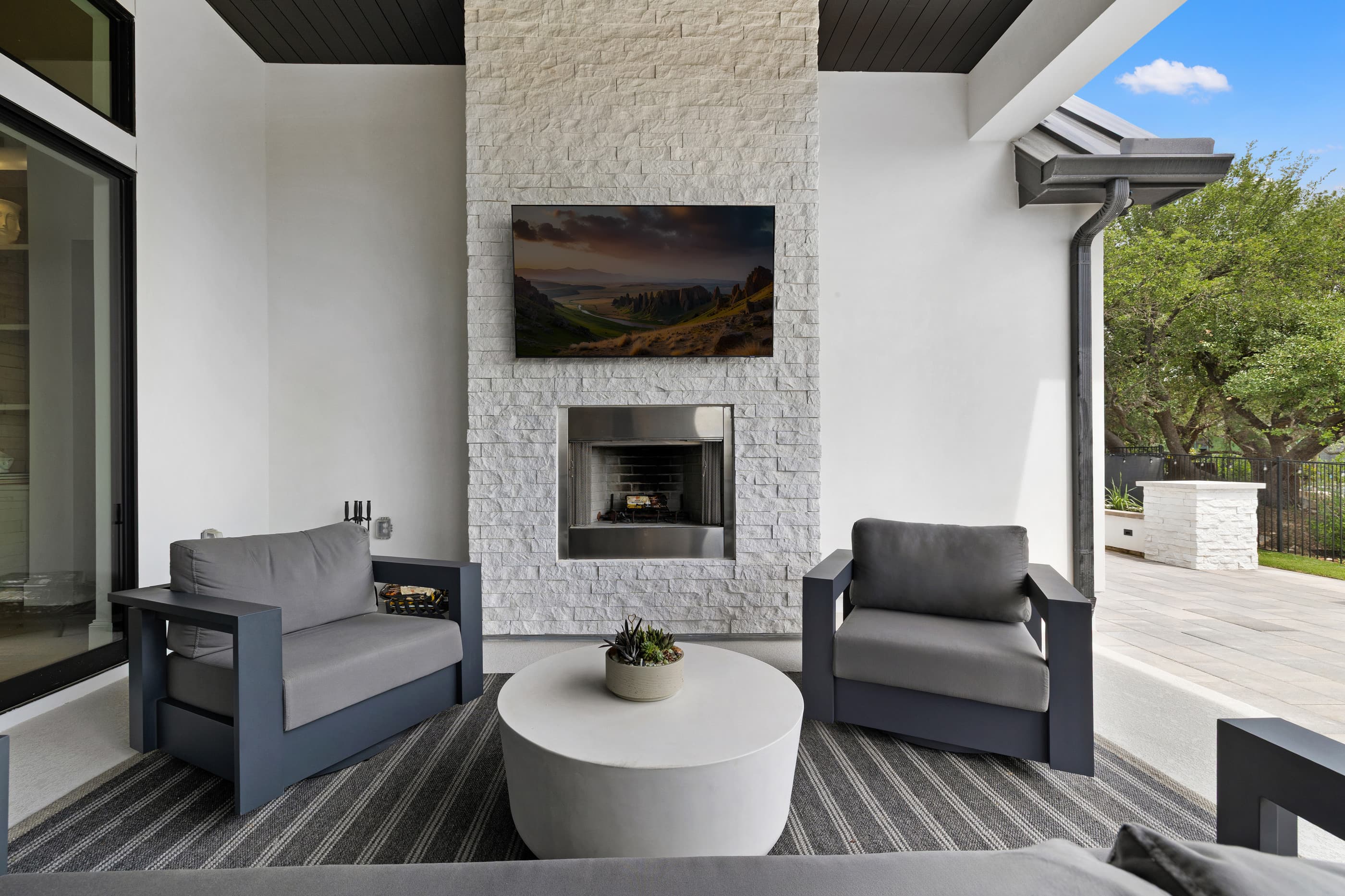 Covered outdoor living room with a stone fireplace, wall-mounted TV and modern lounge chairs