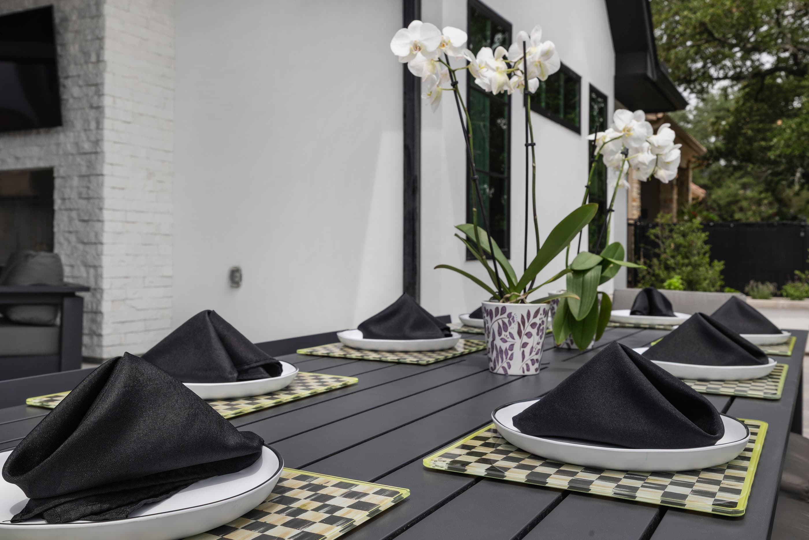 Close-up of a set outdoor dining table with black napkins, orchids and a white stone fireplace behind