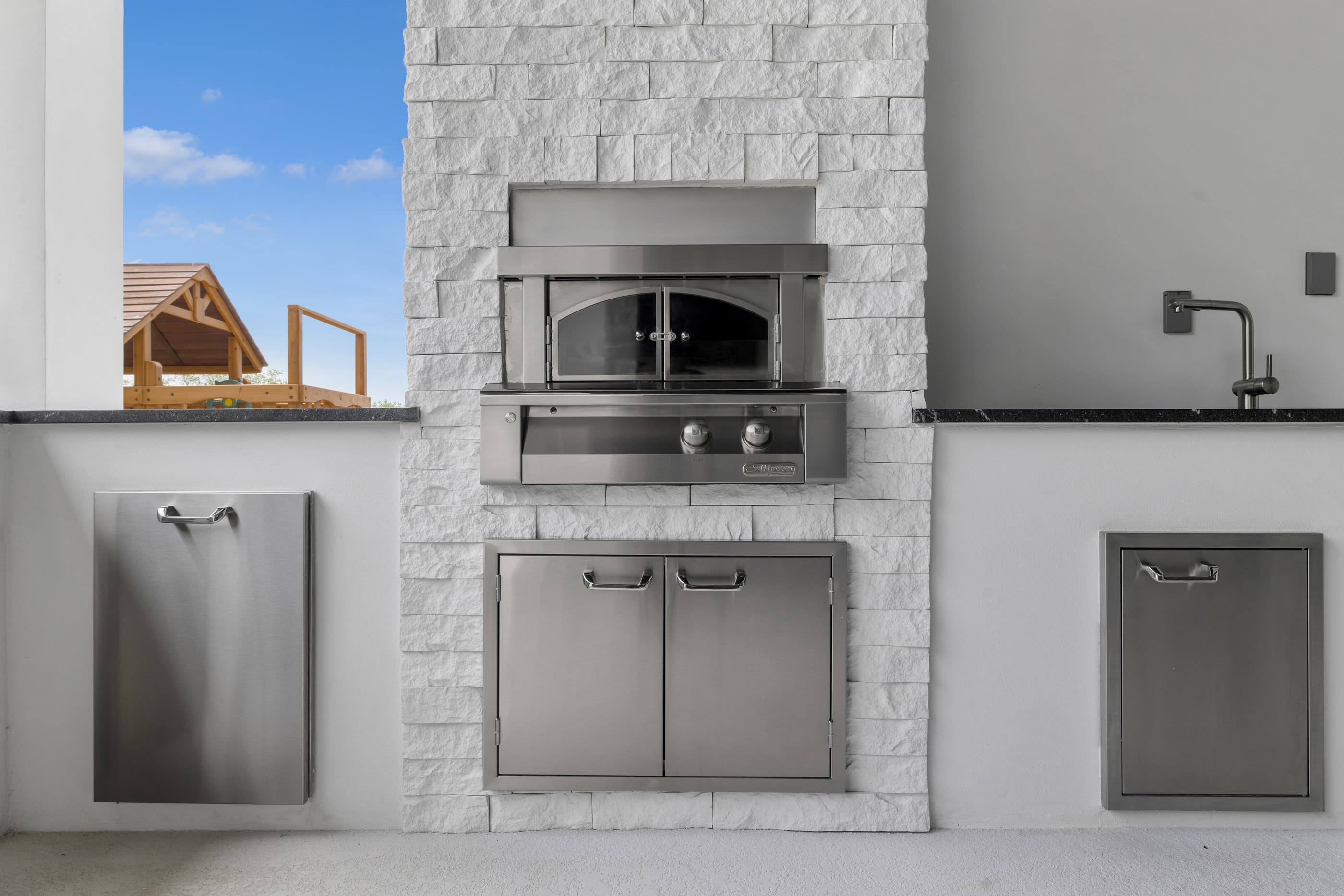 Stone-clad outdoor kitchen with a built-in pizza oven, stainless access doors and bar sink